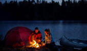 A man and a woman sitting by a bonfire outside their tent at night. Behind them there is a lake and a forest. Beside them there are a canoe.