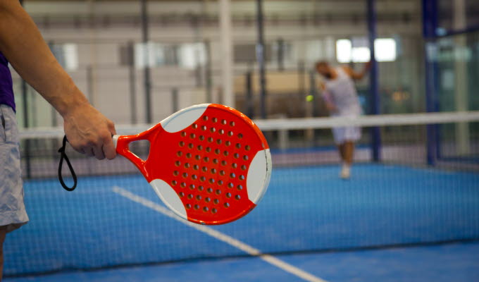 En bild på ett padelrack som någon håller i, i bakgrunden kan man skymta ett nät och en motspelare. 